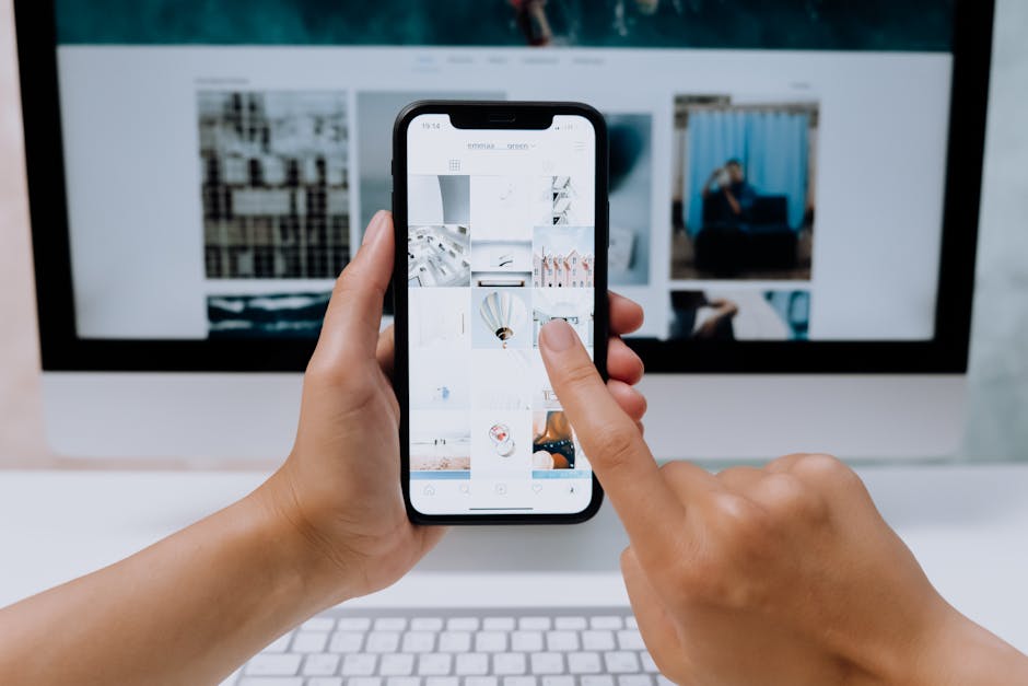 Hands holding a smartphone while browsing a social media app in front of a desktop computer.