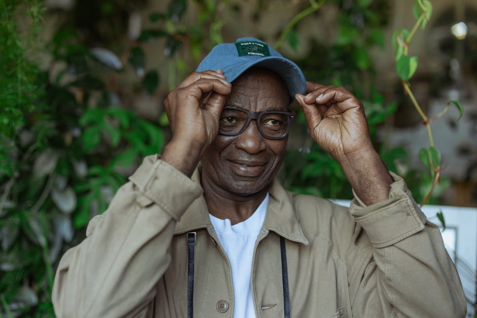 Confident senior man in casual attire adjusting glasses with greenery in the background.