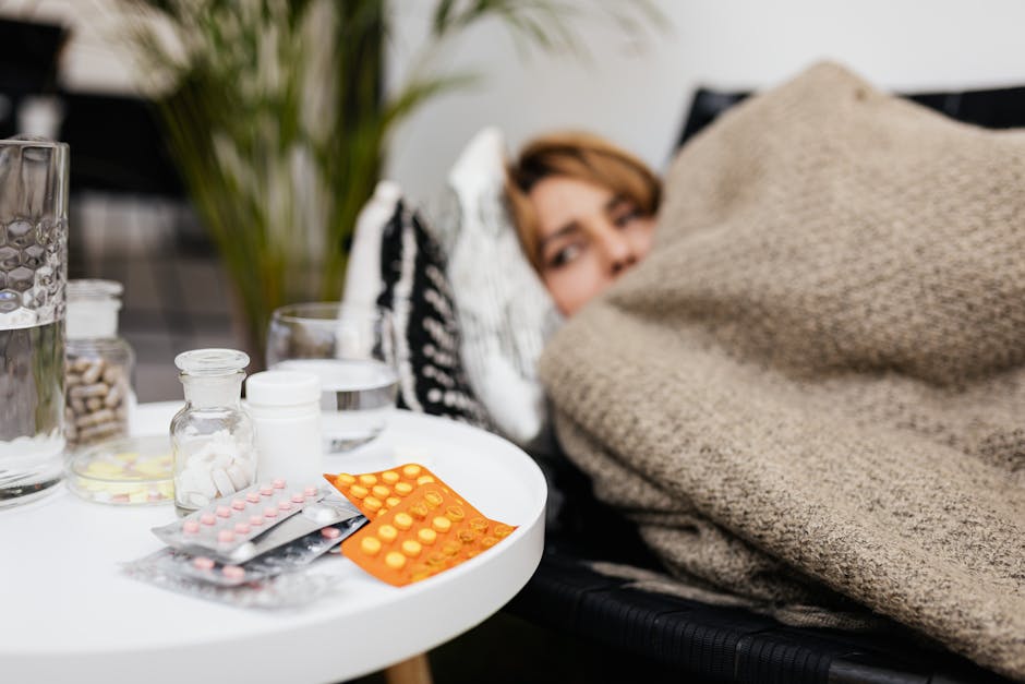 A woman rests under a blanket surrounded by medications on a table, depicting illness and recovery.