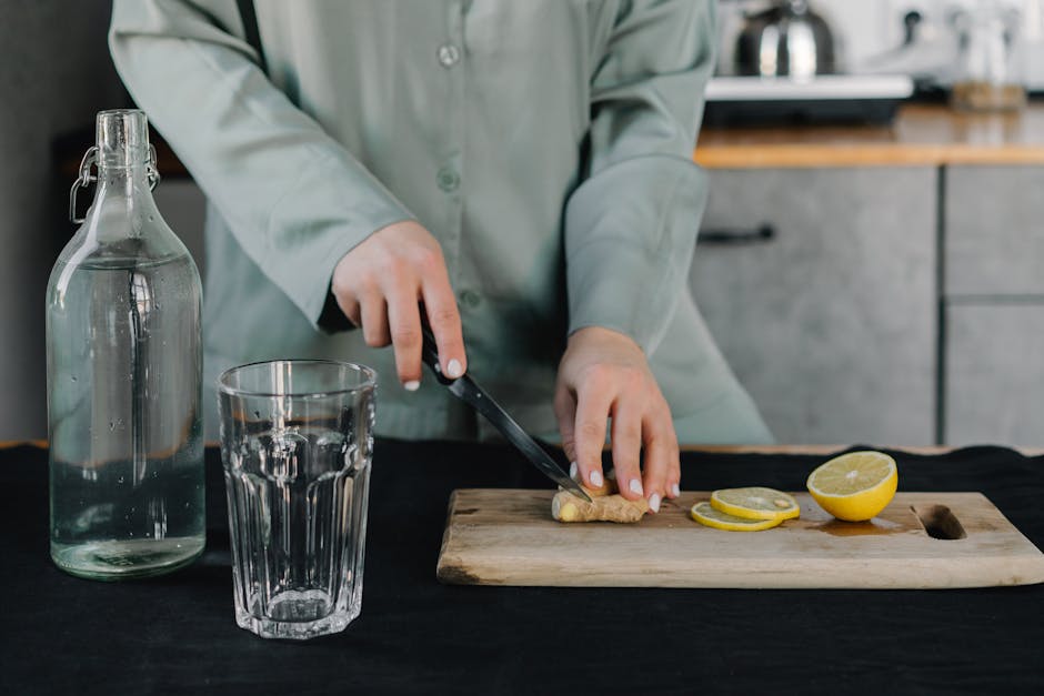 Hands cutting ginger next to lemons, a glass, and water bottle on a wooden board indoors.