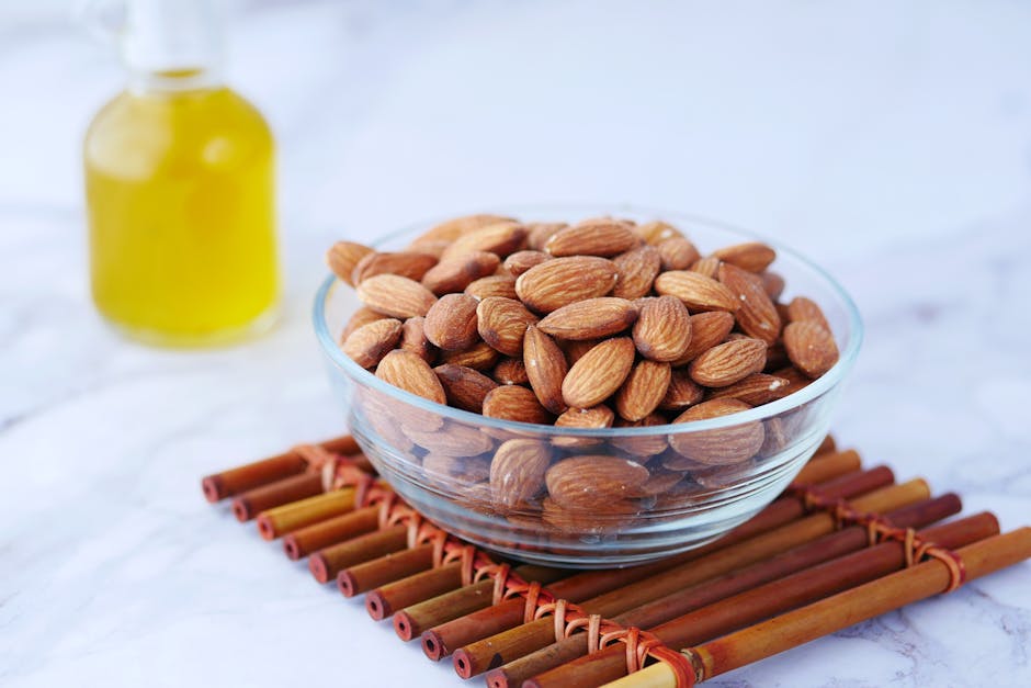 Close-up of almonds in a glass bowl next to a bottle of oil on a bamboo mat.