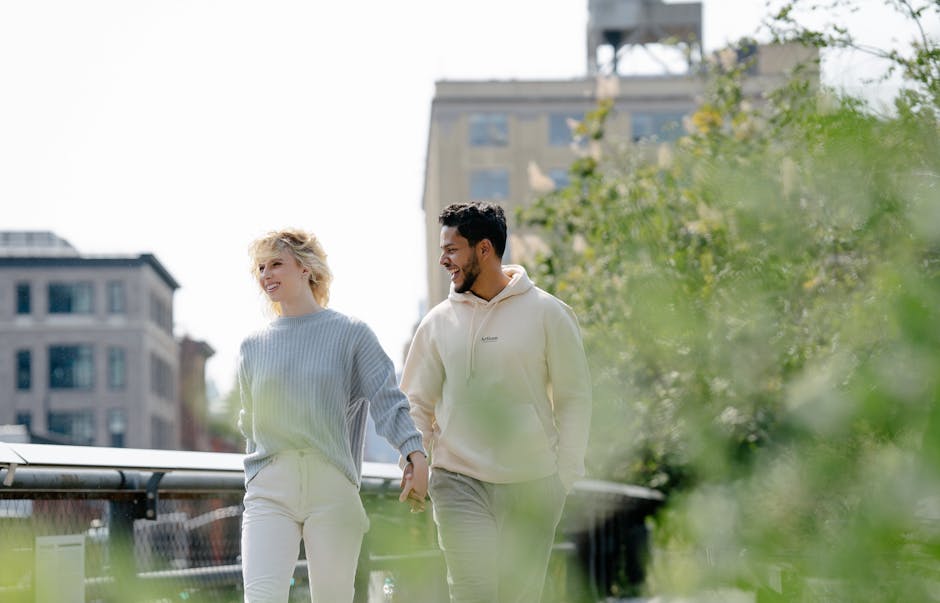 A smiling couple holds hands while walking in an urban park with greenery around.