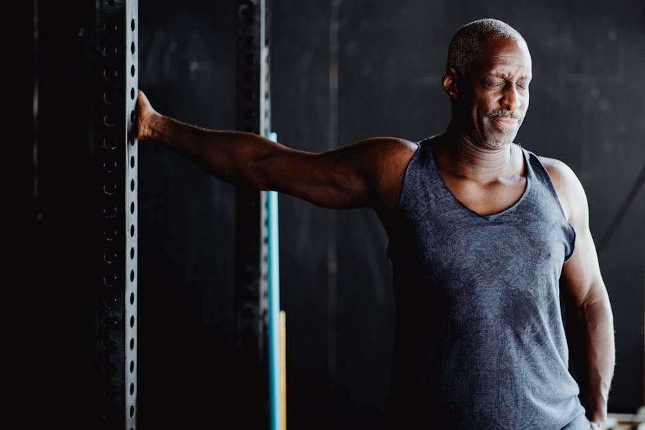 Middle-aged man stretching arm in gym setting while wearing a gray tank top. Fitness and wellness concept.