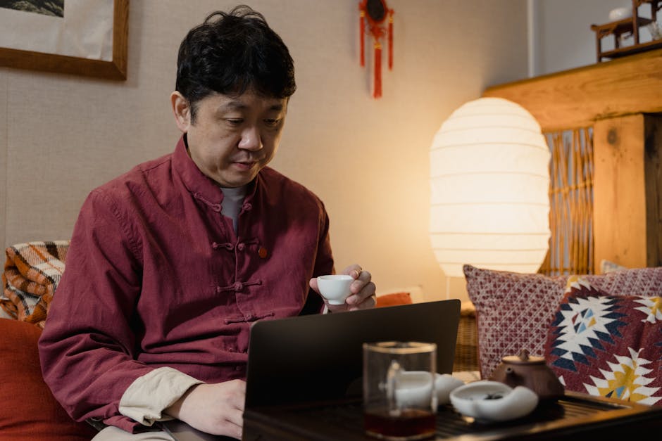Asian man in relaxed indoor setting using a laptop and holding a teacup.