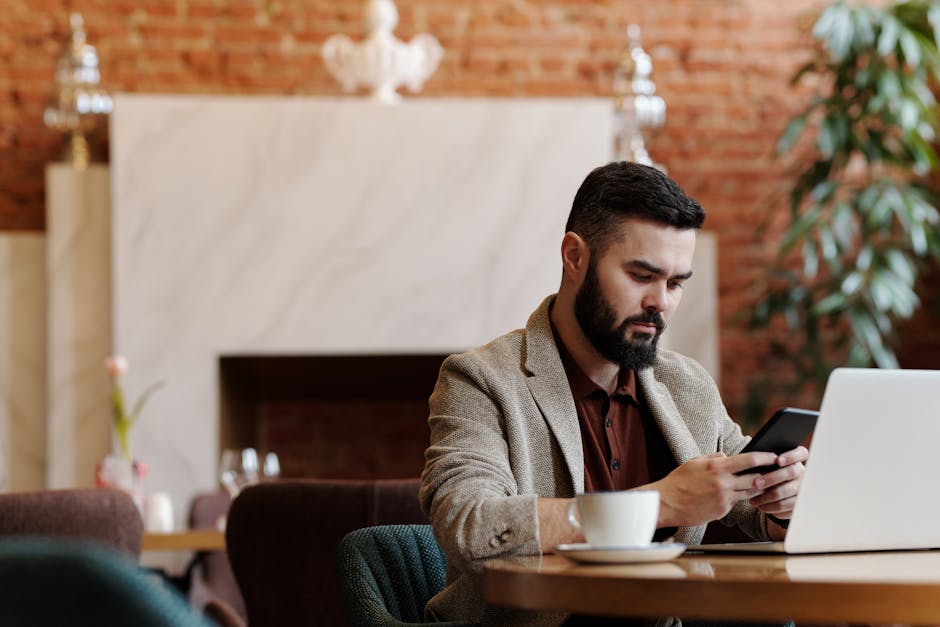 Bearded man using smartphone and laptop for remote work in a trendy coffee shop setting.