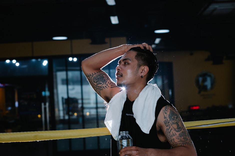 Side view of tired tattooed Latin American male in sportswear with white towel around neck standing in gym with bottle of water during workout